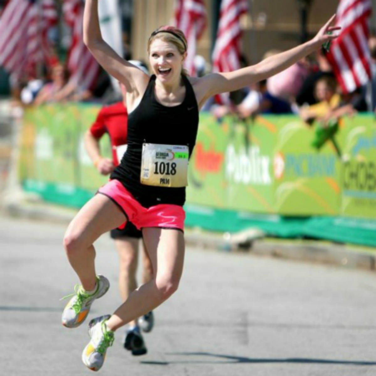 female runner in a black shirt &  pink shorts jumping at the finish line of a race