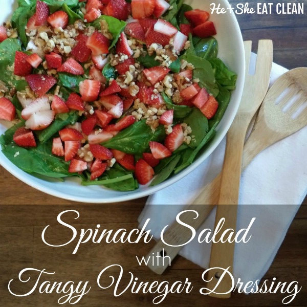 spinach salad topped with strawberries in a white bowl with wooden utensils on the side