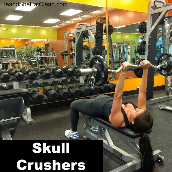 woman lying on a bench doing a skull crushers exercise with arms straight up holding a ez bar