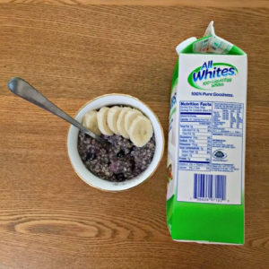small bowl of blueberry oats with sliced banana on top with a spoon. next to the bowl of oats is a carton of allwhite liquid egg whites