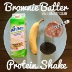 chocolate protein shake in a clear glass with a banana, avocado, and egg white container on the side