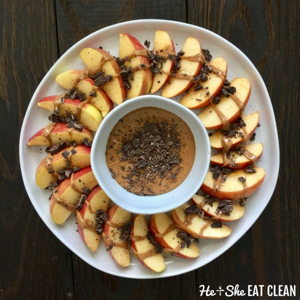 sliced red apple placed in the shape of a wreath with peanut butter in a bowl in the middle and chocolate chunks scattered throughout