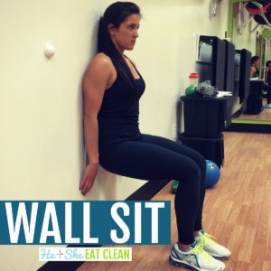 female dressed in black doing a wall sit against a gym wall