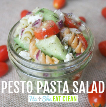 clear mason jar of pasta salad consisting of pasta, cucumbers, tomatoes, red onion, and feta cheese with small tomatoes scattered around the placemat